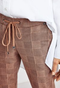Pantalons à carreaux marron avec un motif mélangé, dotés d'une taille à cordon de serrage et d'un tissu texturé. Associés à une chemise blanche visible au niveau de l'ourlet.