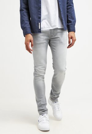 Mand iført lysegrå slim fit jeans, hvide sneakers, hvid t-shirt og en opknappet mørkeblå skjorte, der går fremad på hvid baggrund.