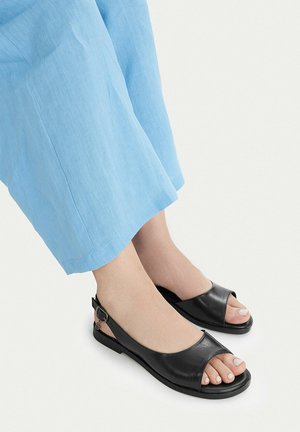 Nohy v černých sandálech s otevřenou špičkou a stahováním za patou, v kombinaci s světle modrými lněnými kalhotami na bílém pozadí.