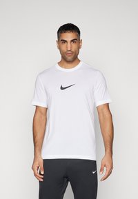 Camiseta blanca de Nike, de manga corta, cuello redondo, con el logo negro de Nike en el pecho, material de algodón suave, combinada con pantalones oscuros.
