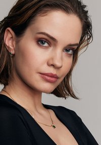 Złoty naszyjnik z zawieszką w kształcie zielonego kamienia szlachetnego. Modelka ubrana w czarną bluzkę, z delikatnymi falami we włosach i naturalnym makijażem.