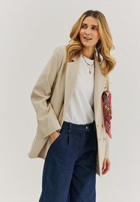 Beige dvouřadový blazer s širokými klopami, v kombinaci s bílým tričkem a tmavě modrými kalhotami s vysokým pasem. Výrazná růžová šála s vzorem jako akcent.