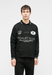 Giovane uomo che indossa una polo nera a maniche lunghe con testo e toppe bianche che recitano "Chalet Club" e pantaloni neri, in piedi su uno sfondo bianco.