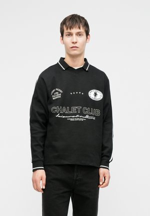 Giovane uomo che indossa una polo nera a maniche lunghe con testo e toppe bianche che recitano "Chalet Club" e pantaloni neri, in piedi su uno sfondo bianco.