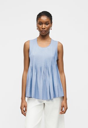 Kobieta z zaplecionymi włosami, nosząca czerwone kolczyki, jasnoniebieski plisowany top bez rękawów i białe spodnie, zwrócona przodem na jednolitym tle.