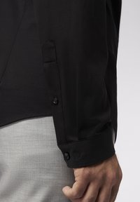 Camisa negra de manga larga con puño puntiagudo, presenta un cierre de botón y tela texturizada. El puño muestra un sutil detalle bordado.