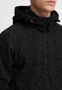 Black fleece jacket with a textured finish, zip closure, stand-up collar, and adjustable hood featuring toggle accents.