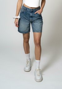 Lysblå denimshorts med en afslappet pasform, der har høj talje, to forlommer og en frynset kant. Kombineret med hvide sneakers.