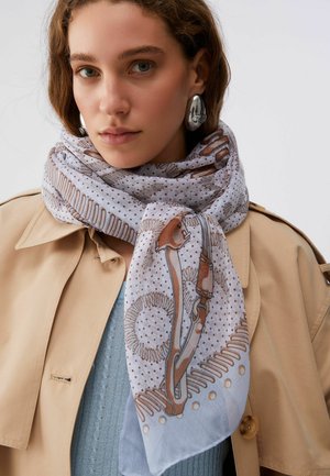 Donna che indossa un trench beige, un maglione azzurro chiaro, una sciarpa decorata con motivi marroni e blu, e grandi orecchini ovali argentati.