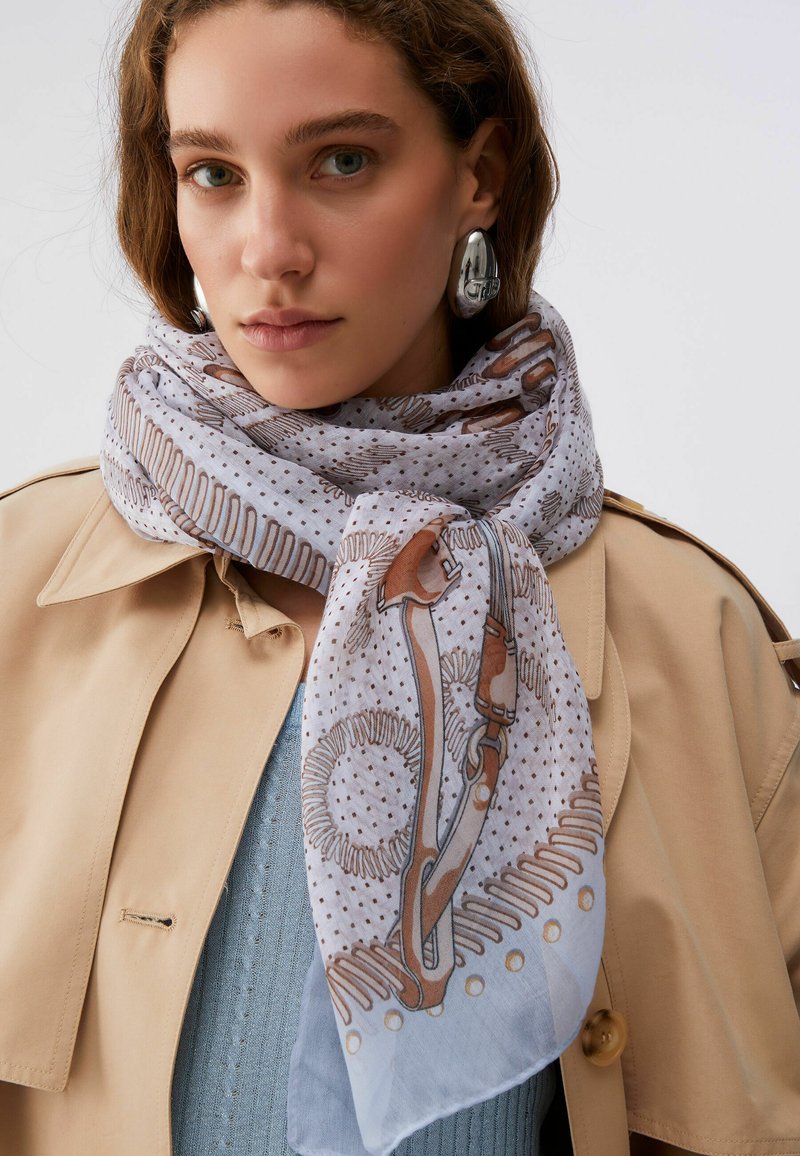 Femme portant un trench-coat beige, un pull bleu clair, une écharpe à motifs avec des dessins marron et bleu, et de grandes boucles d'oreilles ovales argentées.