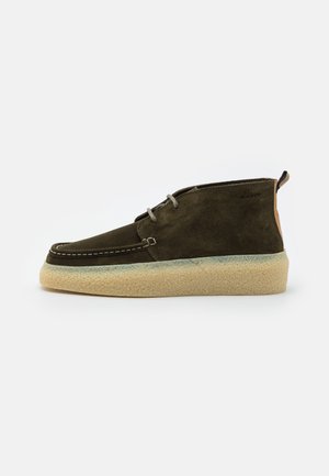Chaussure mi-haute en suède marron avec semelle en crêpe beige, devant lacé et languette de tirage avec branding sur fond blanc.