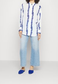Camicia bianca con motivo tie-dye blu, vestibilità rilassata, abbinata a jeans a gamba larga blu chiaro e mule di stoffa blu.