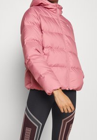 Różowa kurtka puchowa z kapturem, z kwadratowym wzorem i zamkiem błyskawicznym z przodu. Slim fit z elastycznymi mankietami, w połączeniu z czarnymi legginsami.