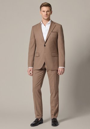 Mężczyzna stoi, ubrany w beżowy garnitur, białą koszulę i czarne mokasyny na jednolitym beżowym tle.
