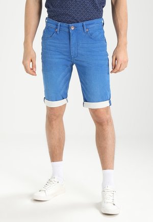 Mand iført opkrogede blå denimshorts, hvide sokker og hvide sneakers, stående mod en ensfarvet lys baggrund.