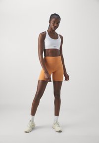 Sujetador deportivo blanco y pantalones cortos de talle alto naranjas. La tela parece suave y elástica. Modelo lleva zapatillas blancas y calcetines de caña corta.