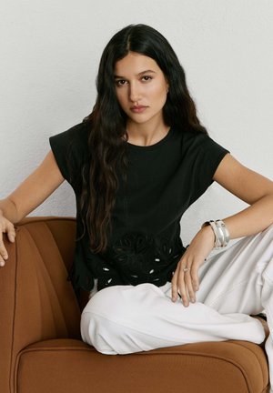 Žena s dugom tamnom kosom, odjevena u crni top s izrezima u obliku cvjetnog uzorka i bijele hlače, sjedi na smeđoj stolici uz bež zid.