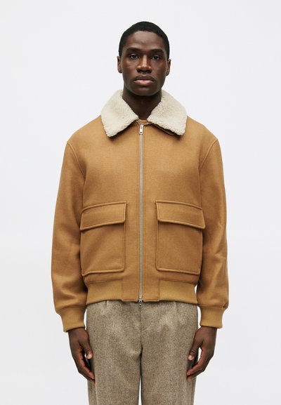 Veste en mélange de laine marron avec un col en fausse fourrure beige, fermeture éclair à l'avant et deux poches avant. Le bas et les poignets côtelés créent une forme structurée.