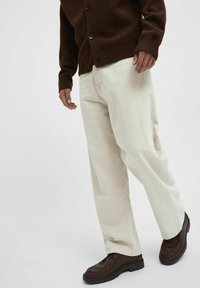 Off-white, loose-fitting pants made of cotton, paired with a dark brown textured cardigan and brown shoes with black soles.