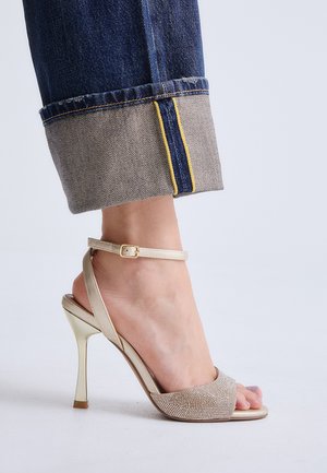 Pie con sandalia de tacón alto beige con correa en el tobillo y piedras de strass, combinado con vaqueros azules remangados sobre fondo claro.