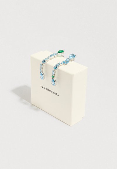 Brincos em tom prateado com várias pedras preciosas azuis e verdes, apresentando várias formas numa caixa de exposição rotulada "Completedworks."