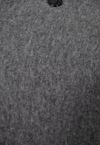 Šedá pletená tkanina s jemnou strukturou má jako výrazný prvek kulatý černý knoflík. Jemné variace v odstínech dávají hloubku.