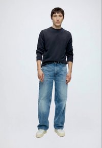 Tânăr în picioare, poartă un tricou negru cu mâneci lungi, blugi largi bleu deschis și adidași albi, pe un fundal simplu și deschis la culoare.