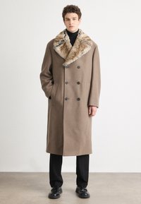 Abrigo de lana marrón con un cuello de piel texturizada, diseño de doble botonadura, botones grandes y bolsillos laterales. Llevado sobre pantalones y zapatos negros.