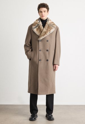 Abrigo de lana marrón con un cuello de piel texturizada, diseño de doble botonadura, botones grandes y bolsillos laterales. Llevado sobre pantalones y zapatos negros.