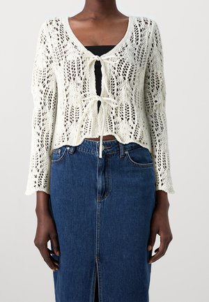 Femme portant un cardigan blanc en maille ajourée avec des liens à l'avant sur un top noir et une jupe en denim bleue taille haute avec une fente devant.