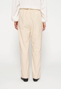 Lichtbeige katoenen broek met een elastische tailleband, rechte pijpen en omgeslagen zoom, gezien van de achterkant. Zwarte schoenen zijn zichtbaar.