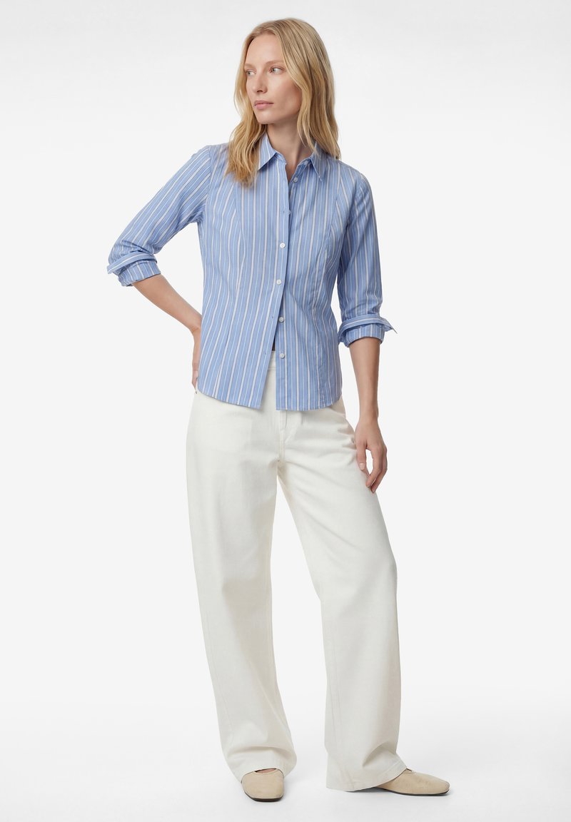 Mujer con una camisa de rayas azules abotonada, con las mangas remangadas, pantalones blancos de pierna ancha y zapatos beige, de pie con una mano en la cadera.