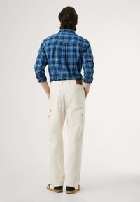 Blauwe geruite shirt met lange mouwen, witte cargobroek met zakken en zwarte instap schoenen met tan kleurige zolen. Van achter gezien.