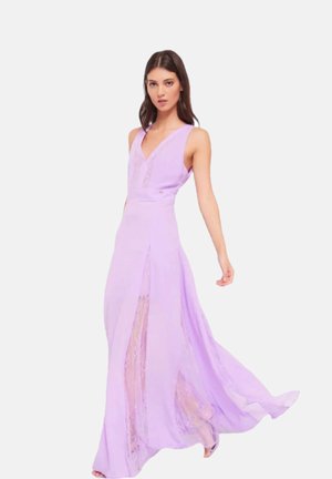 Donna che indossa un abito lavanda senza maniche, fluido e con pannelli di pizzo, mentre cammina con un braccio esteso all'indietro su uno sfondo bianco semplice.