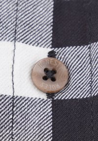 Ruudullinen kangas, jossa on musta ja valkoinen neliö, jonka päällä on ruskea, nelireikäinen nappi, jossa lukee "HONEST" ja "PULSES", ja musta lanka näkyy.