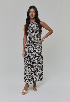 Maxikleid in einem schwarzen und cremefarbenen Zebramuster mit figurbetontem Mieder, ausgestelltem Rock und Seitentaschen; kombiniert mit goldenen Mules mit offenem Zehenbereich.