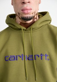 Felpa con cappuccio verde oliva, dotata di tasca a kangaroo, con logo "carhartt" ricamato in viola, doppio laccio e una consistenza morbida.