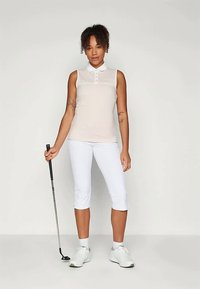 Žena v bezrukávovom béžovom polotričku a bielych kapri nohaviciach, ktorá drží golfovú putterku a stojí pred jednoduchým svetlým pozadím.