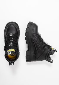 Bottes en cuir noir épaisses avec semelles crantées épaisses, languettes de traction de marque et détails jaunes, vues de dessus et de côté sur fond blanc.