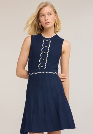 Blondynka w granatowej sukience bez rękawów z białym zygzakowatym wykończeniem i złotymi guzikami, stojąca na jednolitym tle.