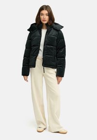 Doudoune noire avec capuche, présentant un design matelassé et une fermeture éclair. Associée à un pantalon large crème et des chaussures beiges.