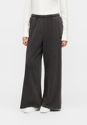 Persoon draagt wijde donkergrijze joggingbroek met elastische taille, wit shirt met lange mouwen en witte sneakers, staand voor een effen achtergrond.