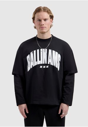 Jonge man met blond haar die een zwarte lange mouwen overhemd met lagen draagt, met grote witte tekst "BALLINAMS XXX" aan de voorkant en een zilveren ketting.