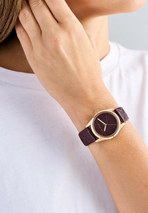 Round watch with a dark purple leather strap, gold bezel, and textured dark purple face featuring simple hour markers and hands.