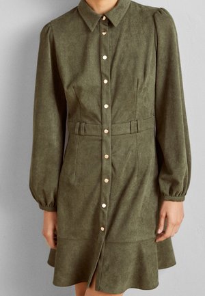 Frau trägt ein olivgrünes, knöpfbares Kleid mit langen Ärmeln, Kragen und tailliertem Gürtel, steht vor einem schlichten Hintergrund.