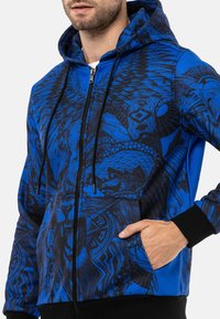 Blaue Zip-Up-Hoodie mit schwarzen grafischen Mustern, ausgestattet mit einer Kapuze mit Kordelzug, Fronttaschen und schwarzen gerippten Bündchen und Saum.