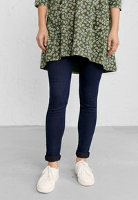 Zelena tunika s uzorkom lišća, uparena s tamnoplave uske traperice i bijelim tenisicama. Tunika ima zakrivljeni rub i rukave do tri četvrtine.