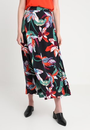 Femme portant une jupe midi noire avec un grand imprimé floral coloré, un haut rouge et des mules noires à talons, debout sur un fond blanc.