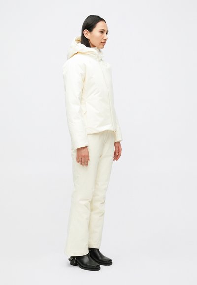 Krém színű, kapucnis, cipzáras szigetelt dzseki, hozzáillő nadrággal párosítva. A modell fekete csizmát visel. Egységesen sima textúra.