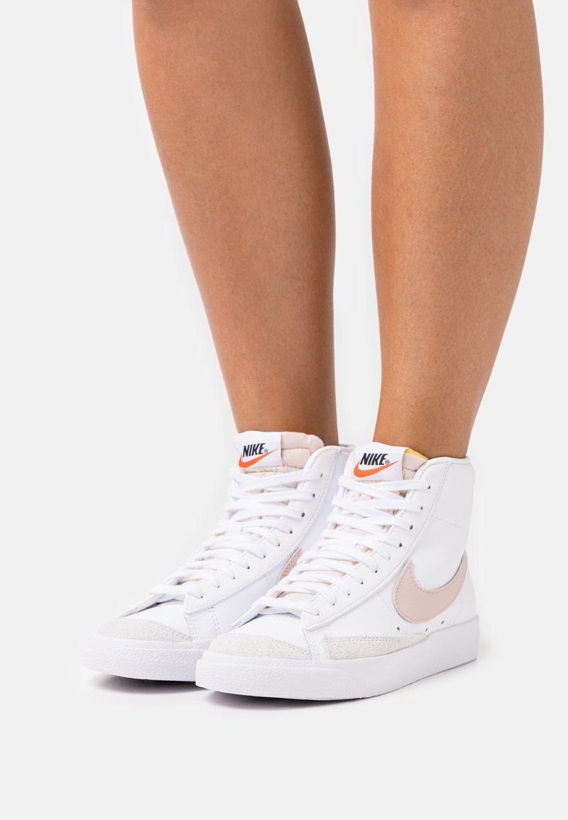 Zapatillas altas blancas con acentos rosas, material de cuero y textil, suela de goma texturizada y distintivo logo Swoosh en el lateral.